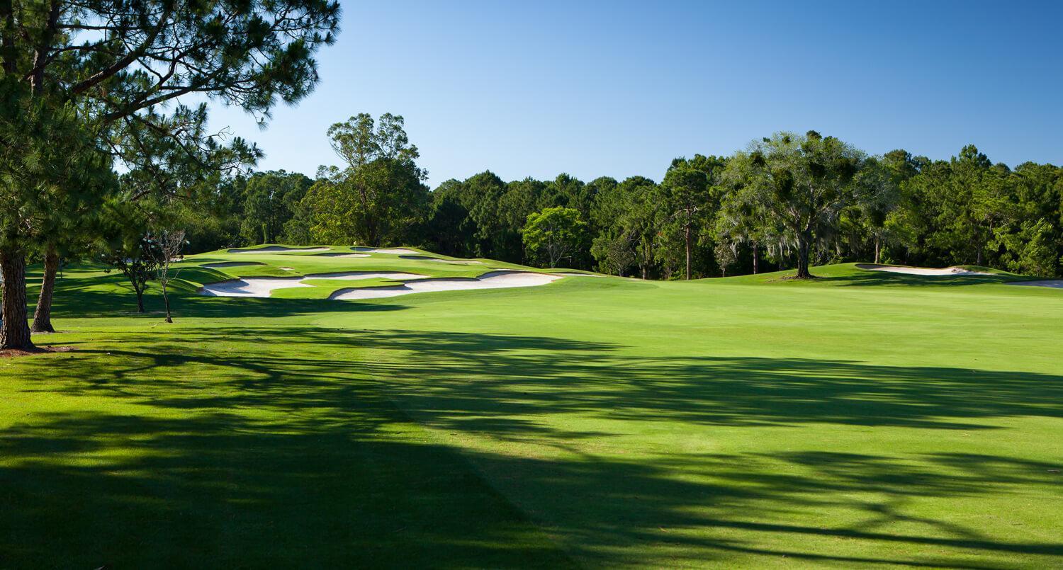 Wide fairway leads to a green littered with sand bunkers under clear blue skies