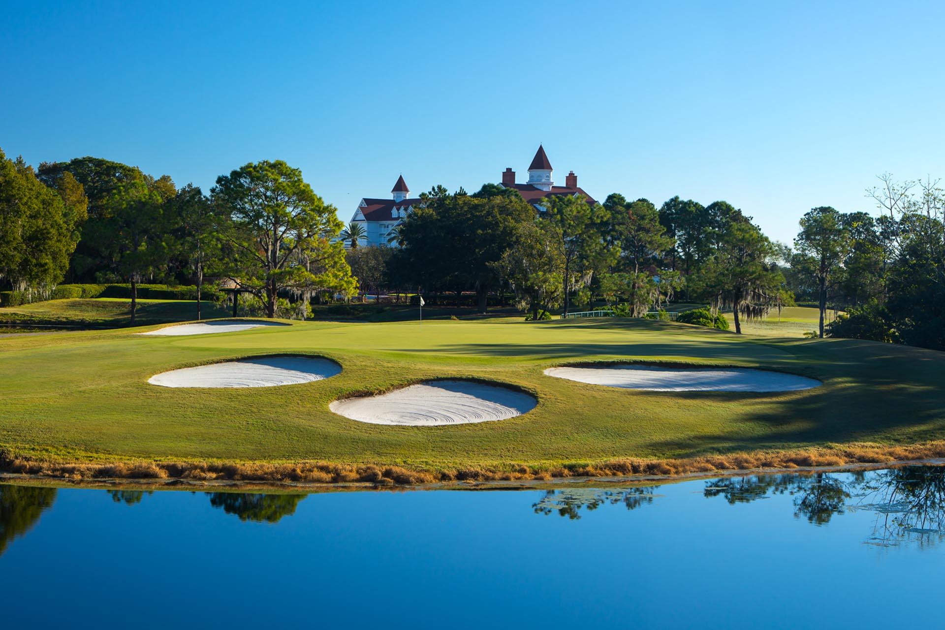 A smooth green surrounded by sand bunkers next to a water hazard with the clubhouse towering over the trees