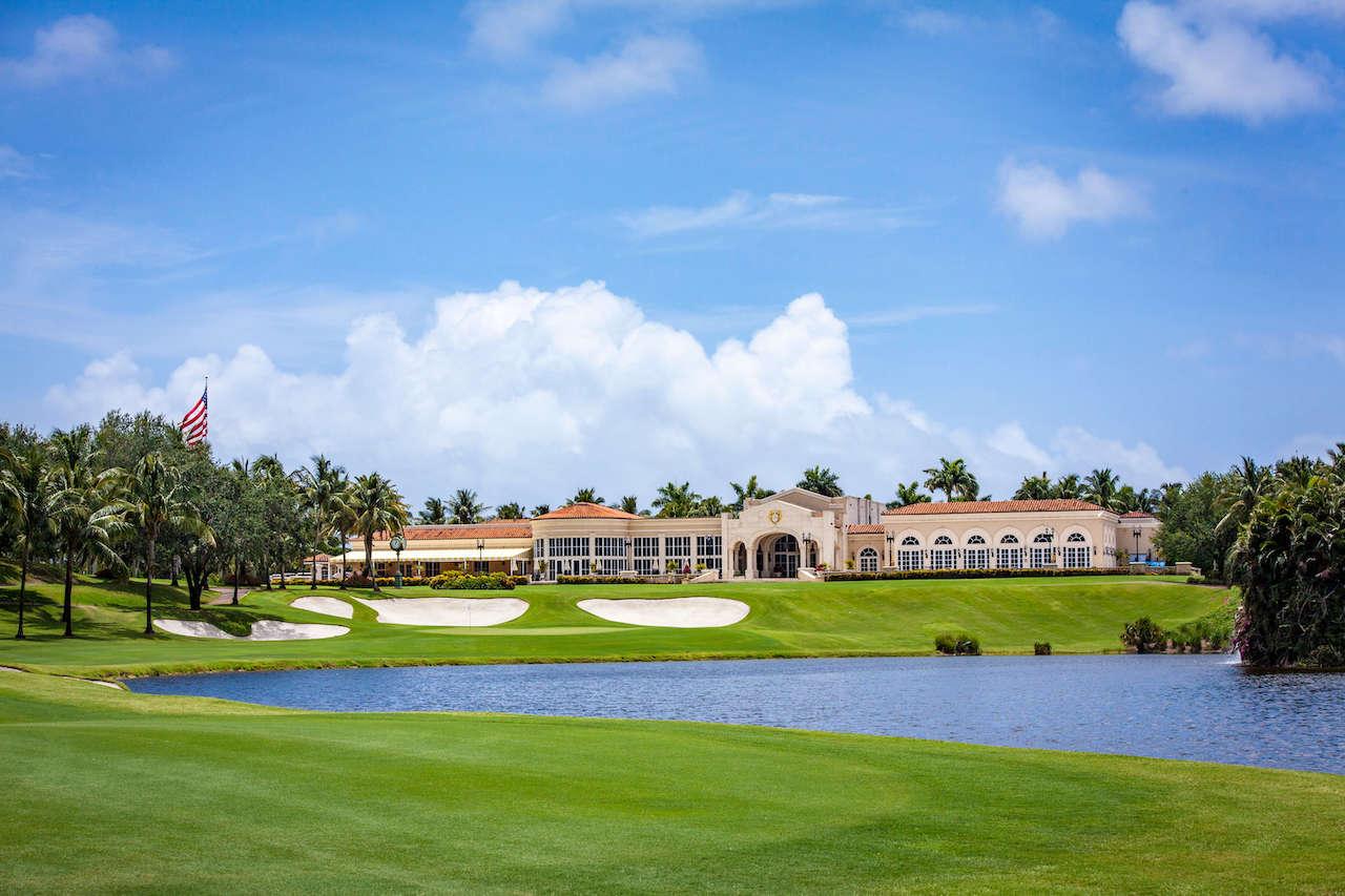 A luxurious golf clubhouse stands tall behind pristine bunkers and a calm water feature under a bright blue sky.