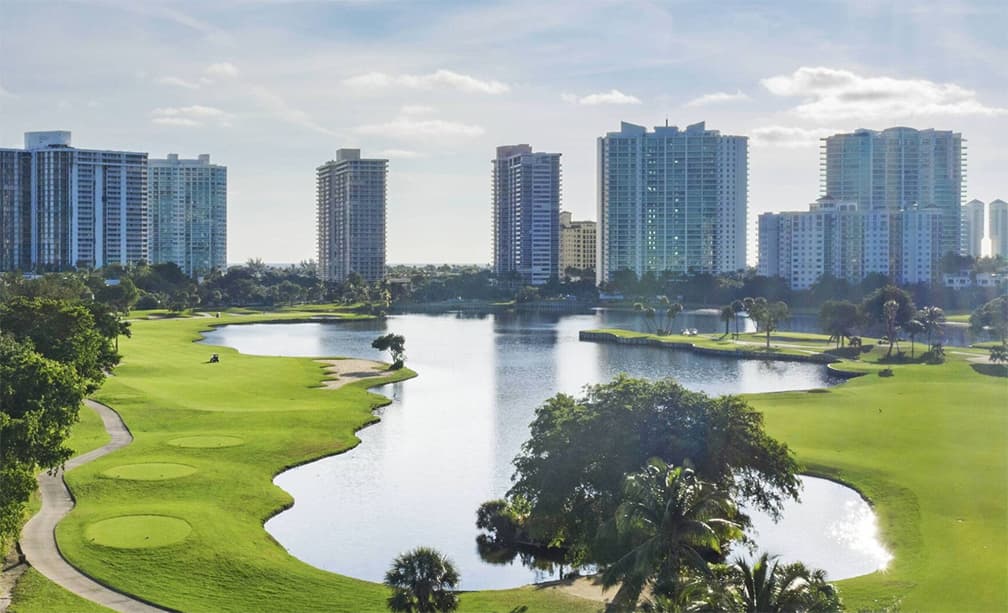 A scenic golf course alongside a reflective lake, with high-rise buildings towering in the distance.
