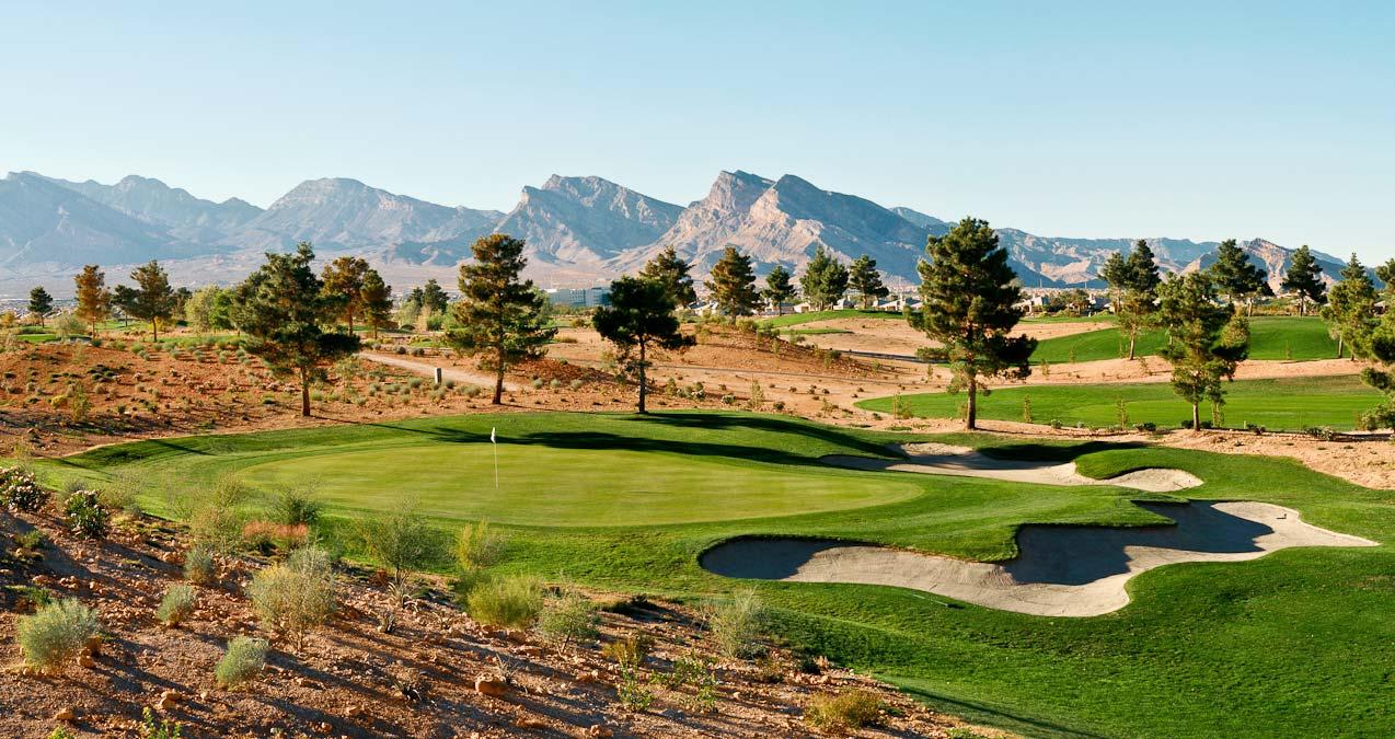 A smooth green surrounded by uniquely shaped sand bunkers littered with trees under blue skies with mountain views in the distance