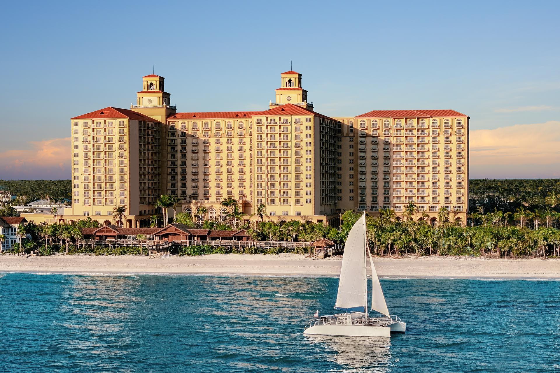 A boat sailing down the shore with The Ritz Carlton Naples grand building towering over the beach