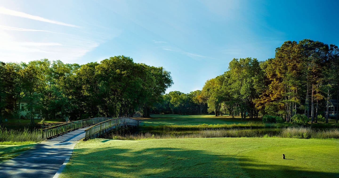A charming wooden bridge leads to a fairway surrounded by lush trees.