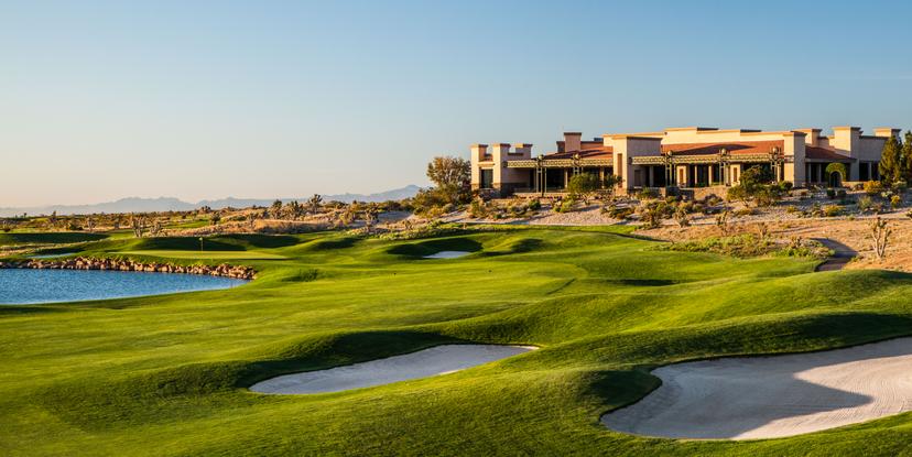 A golf course with sand bunkers and a lakeside view near a desert clubhouse.