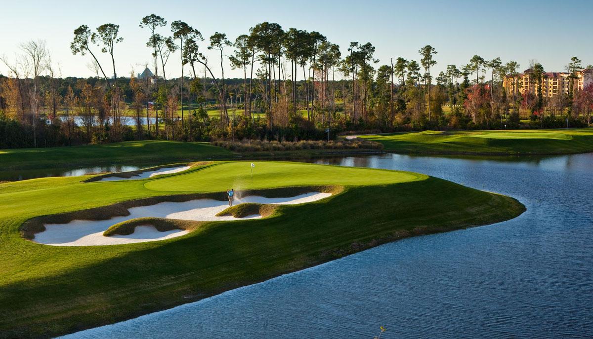 A golfer plays from a bunker on a lush green surrounded by water and pine trees.
