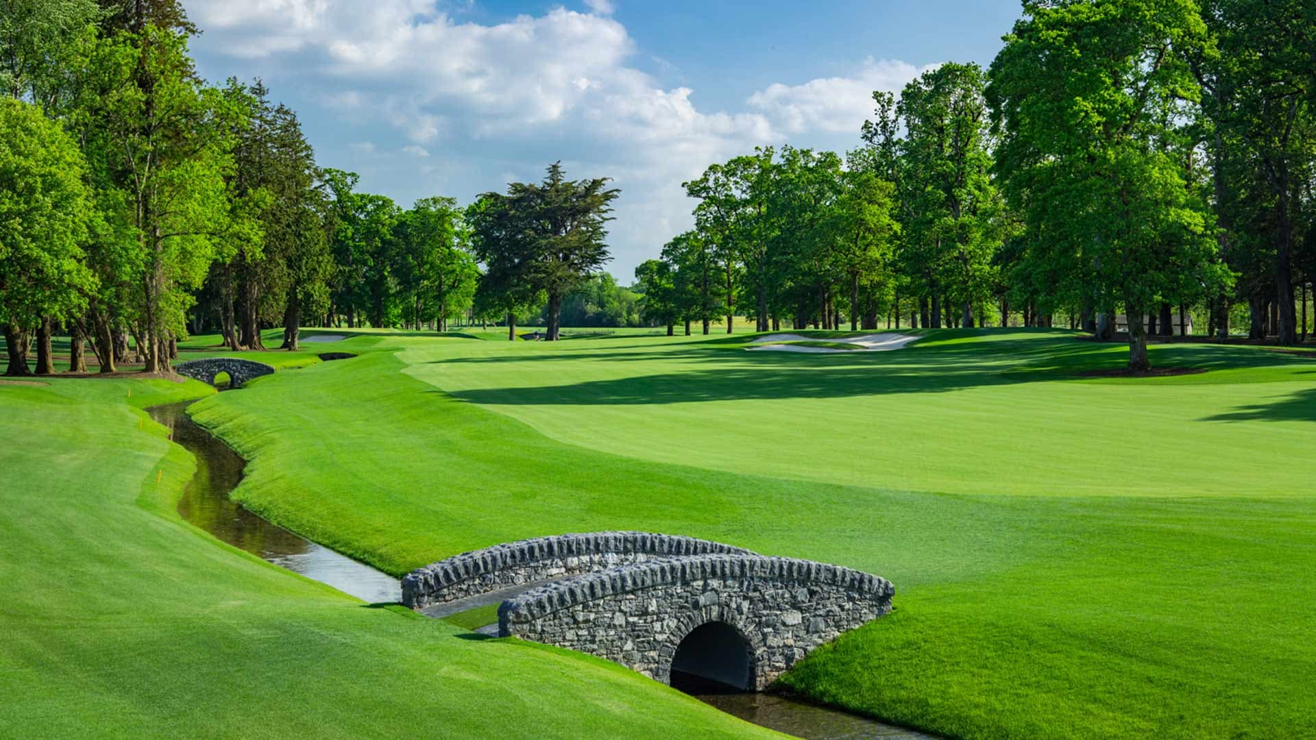 A stone bridge crosses a narrow stream surrounded by lush fairways and tall trees on the golf course.