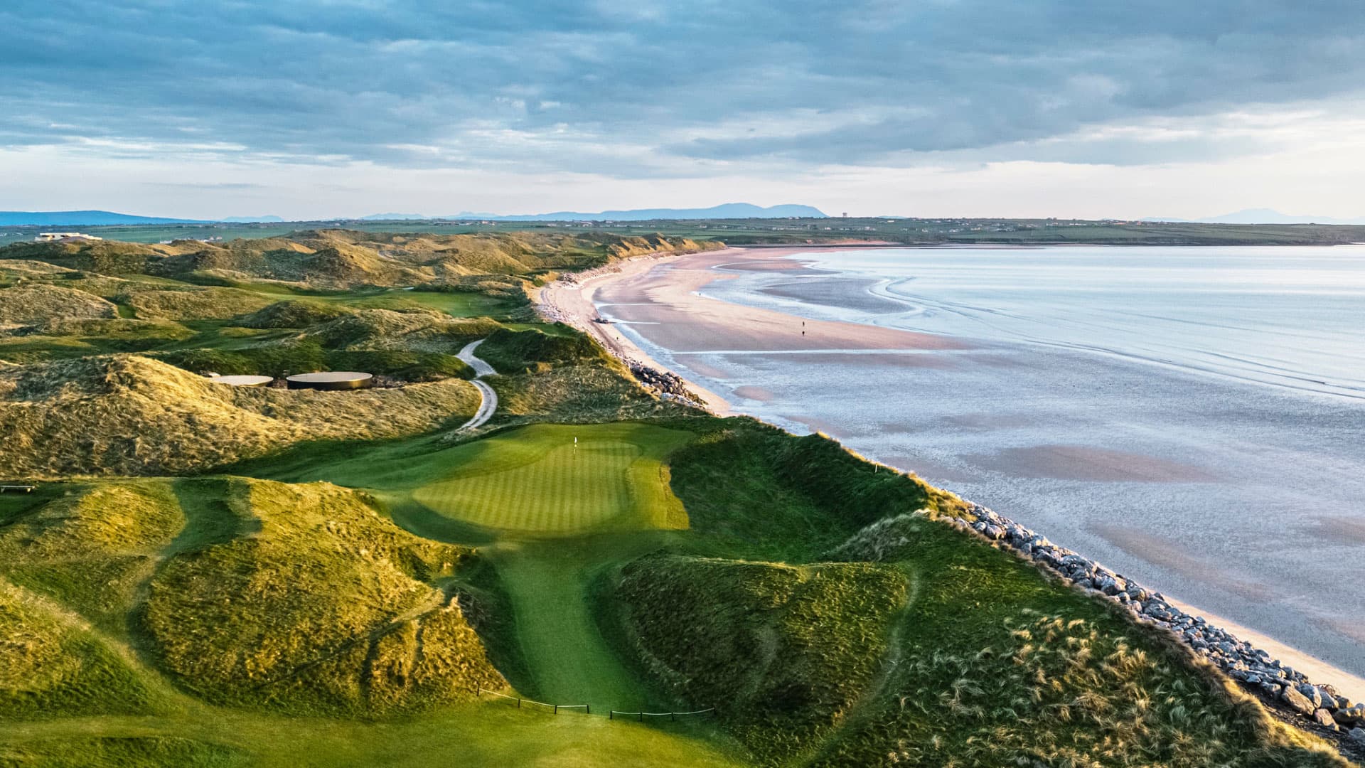 A dramatic aerial view of the course with rolling dunes leading down to a vast sandy beach.