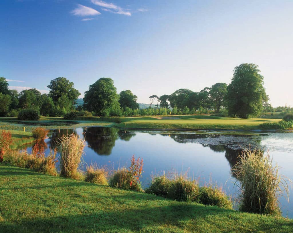 A serene green set beside a reflective water hazard, surrounded by lush trees and vibrant foliage.