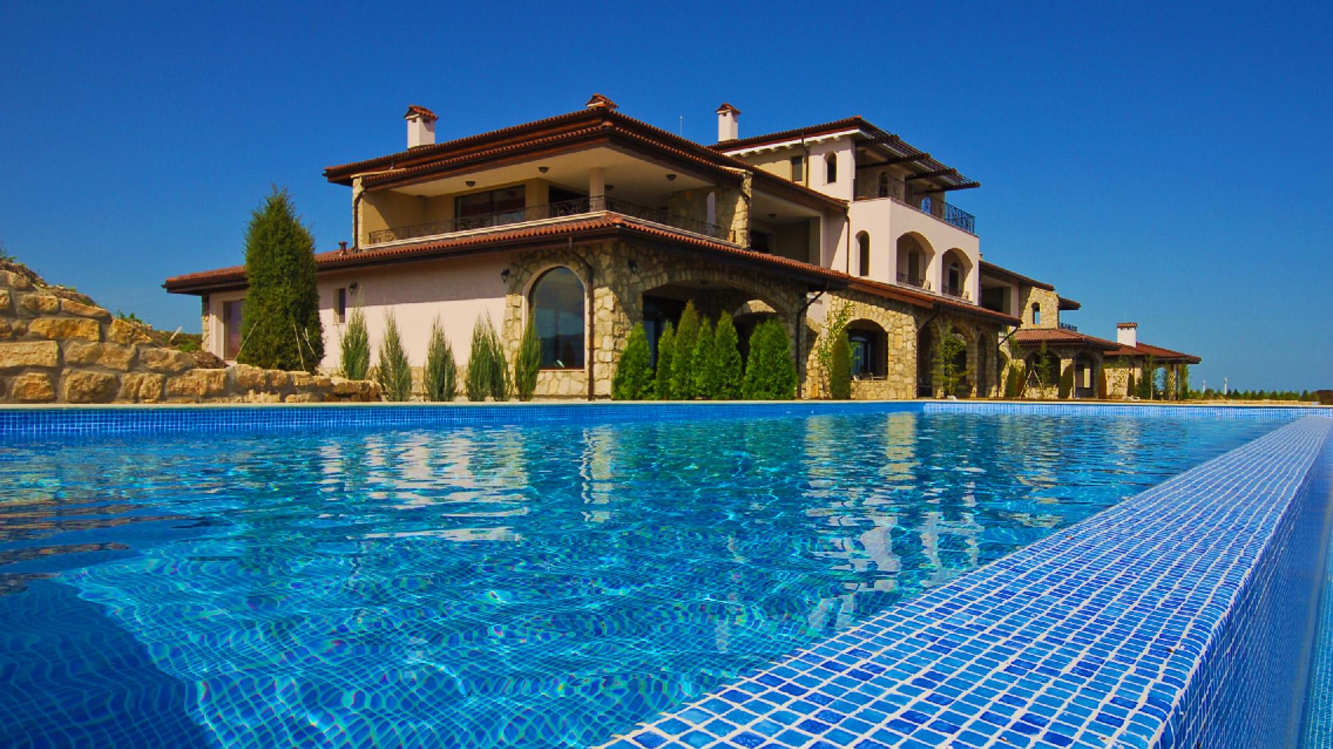 Exterior view of a large three floor villa surrounded by trees leading to a swimming pool