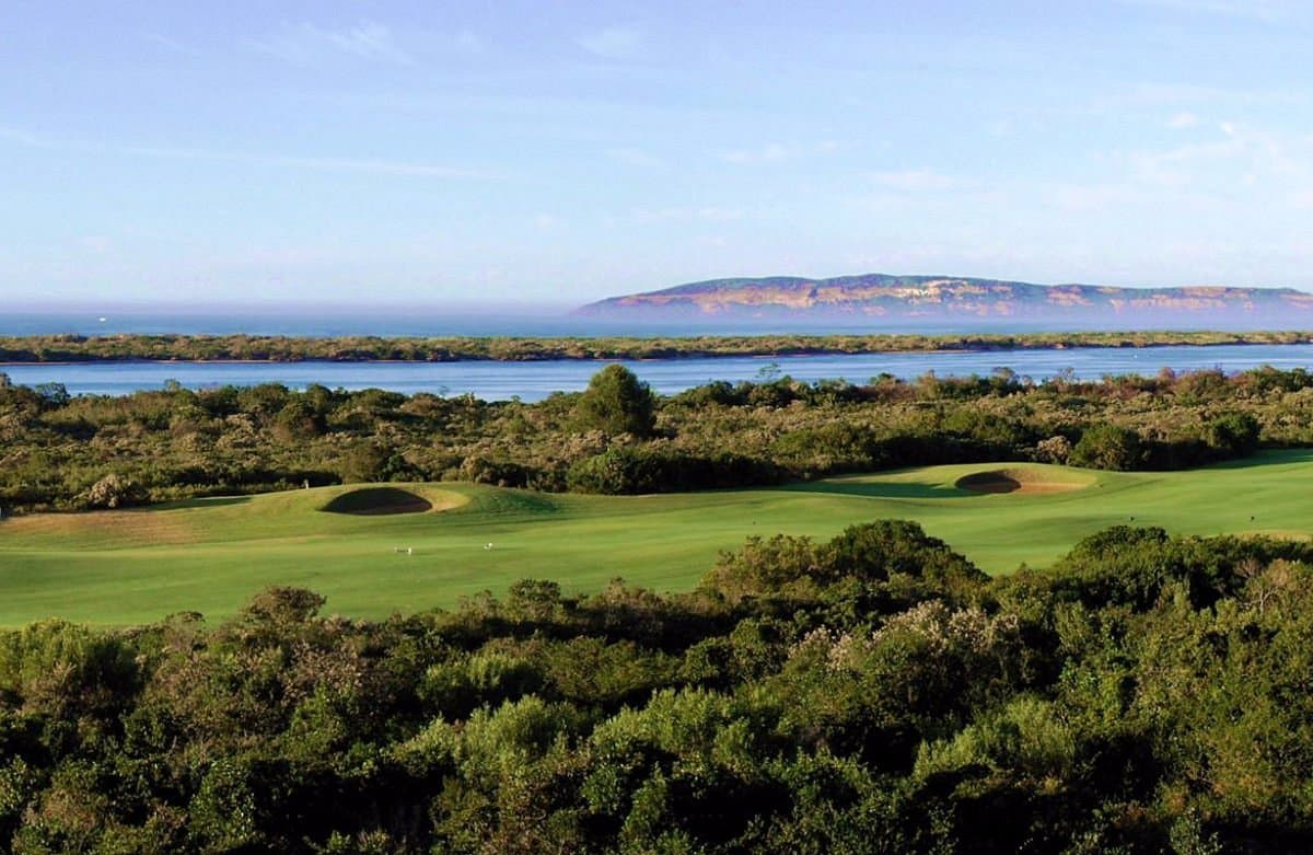 A lush green fairway stretching towards a blue river and distant cliffs.