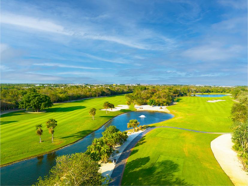 A wide fairway winds alongside a sparkling water feature with palm trees and sand bunkers under a clear blue sky.