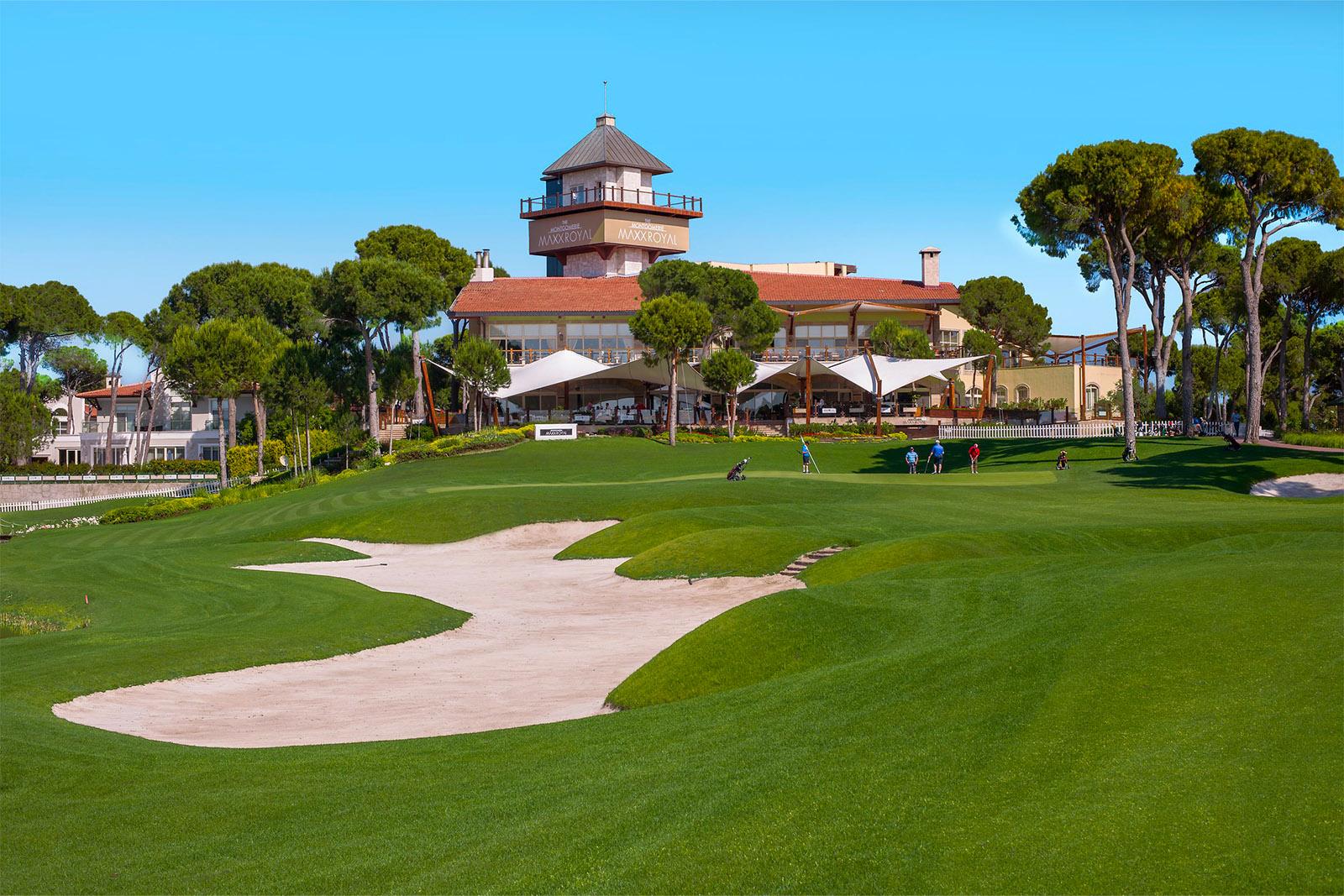 Bunker in the foreground with four players on the green and the clubhouse in the background
