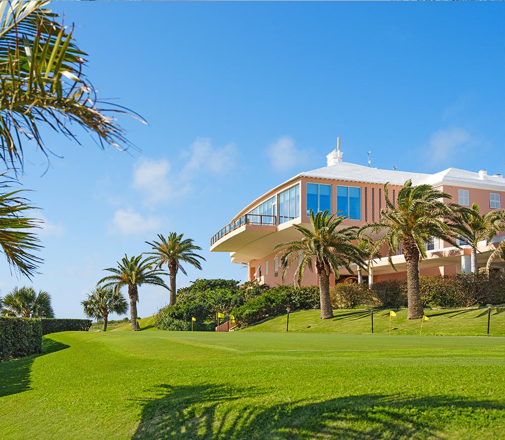 A charming clubhouse with palm trees sits atop a lush green hill under a bright blue sky.