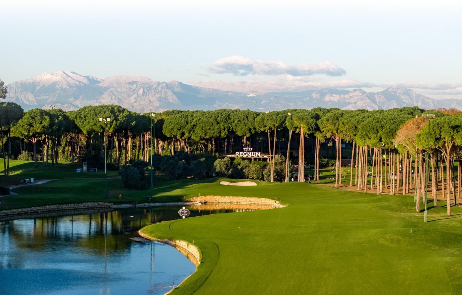 Fairway with water on the left and trees on the right winding towards the green with the clubhouse behind