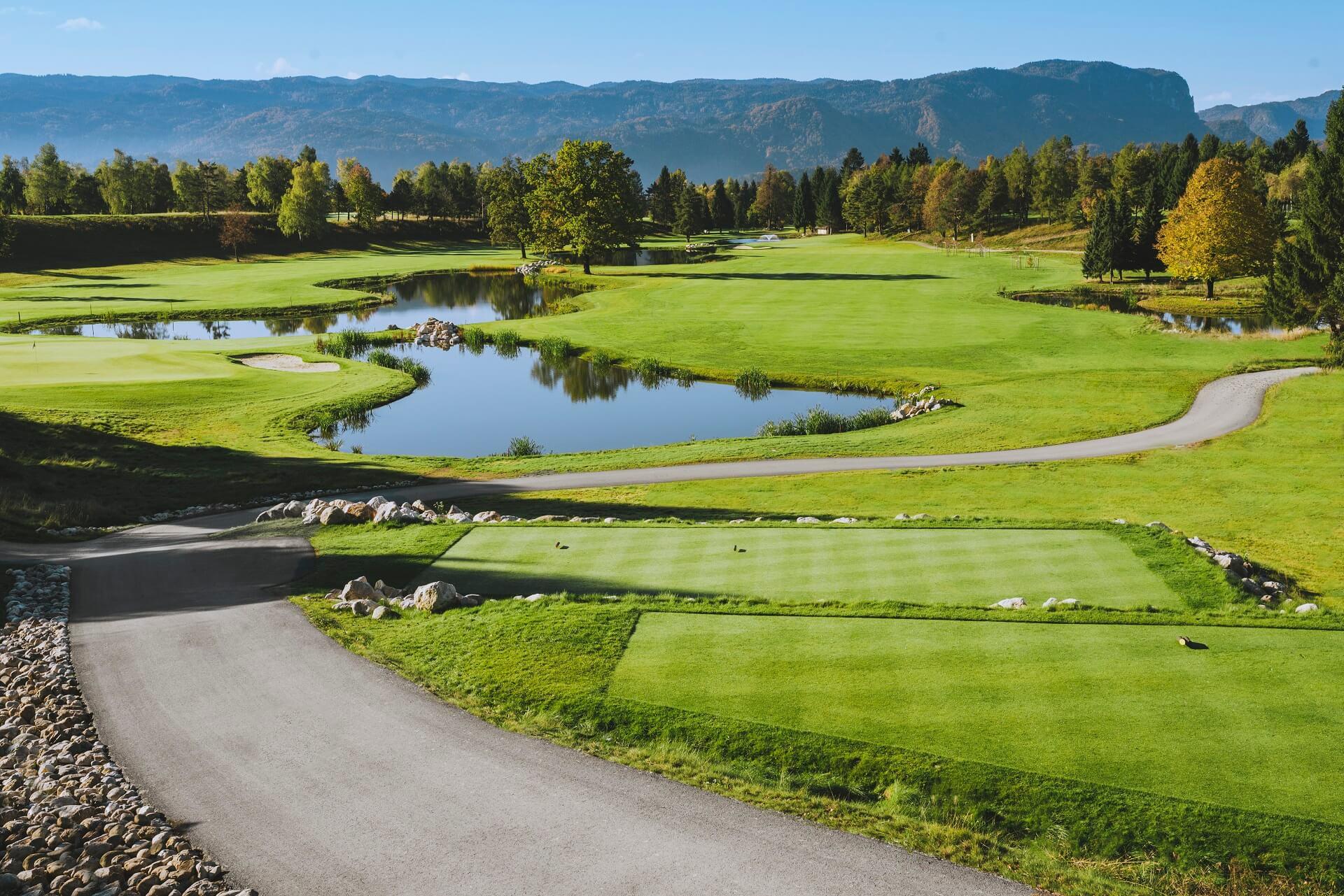 A scenic golf course with a winding path and water features surrounded by mountains.