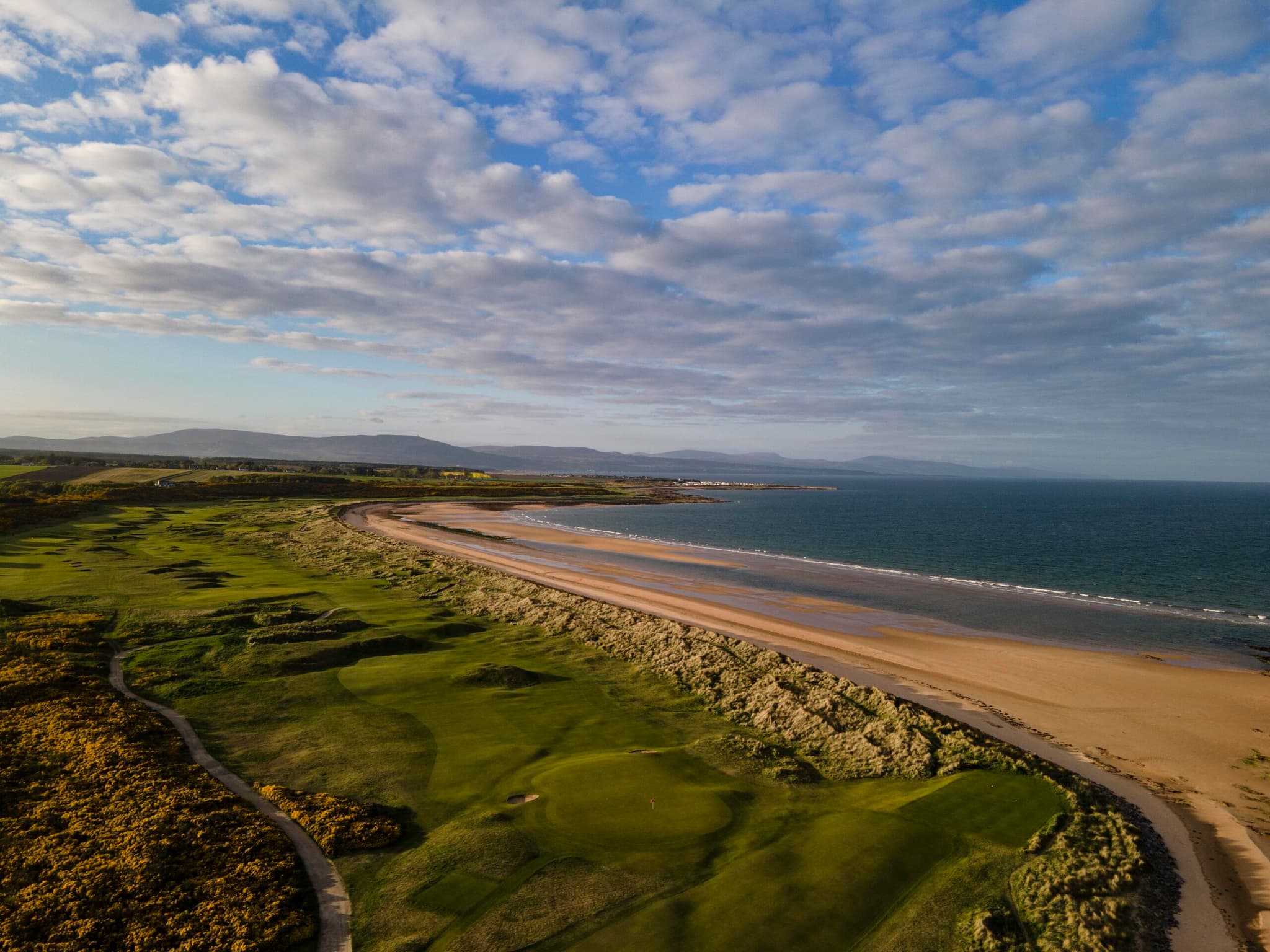 A golf course winds along a sandy coastline under blue skies.