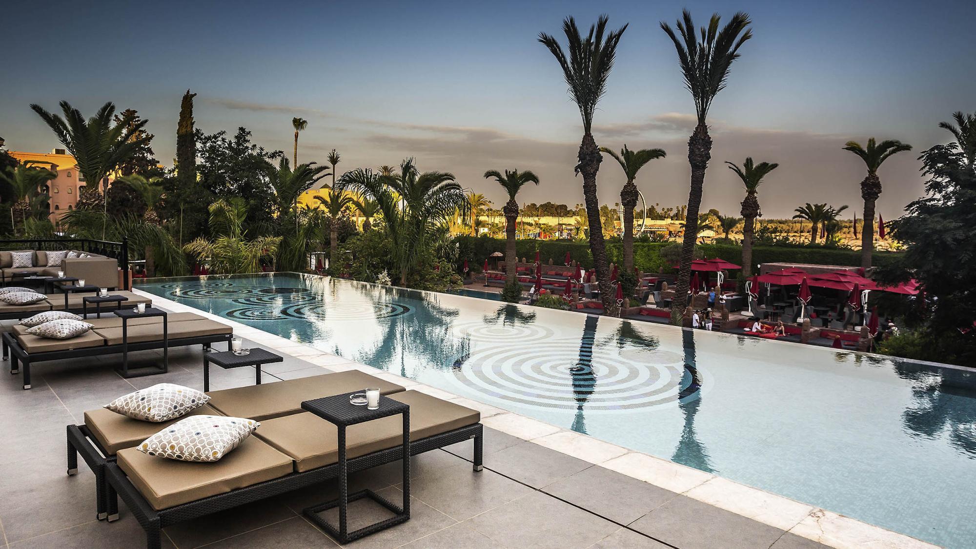 An outdoor swimming pool with a design at the bottom sandwiched between palm trees and sunbeds
