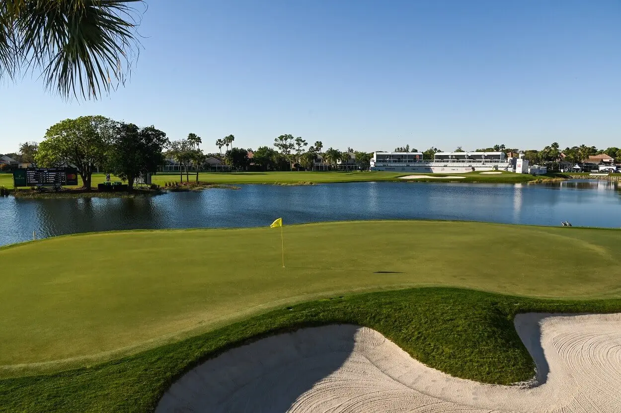 A golf green located near a pond with grandstands and a tournament scoreboard in the background, suggesting a championship course.