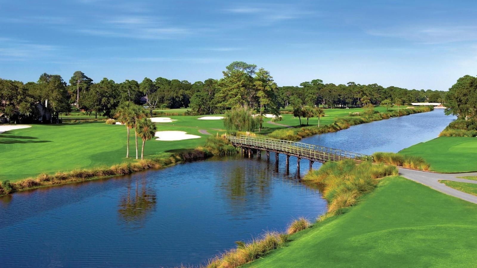 A wooden bridge crosses a river on a sunny golf course.
