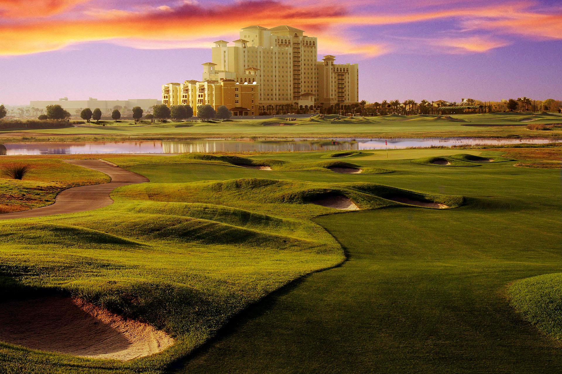 Rolling fairways with deep bunkers lead toward a grand hotel backdrop at dusk.