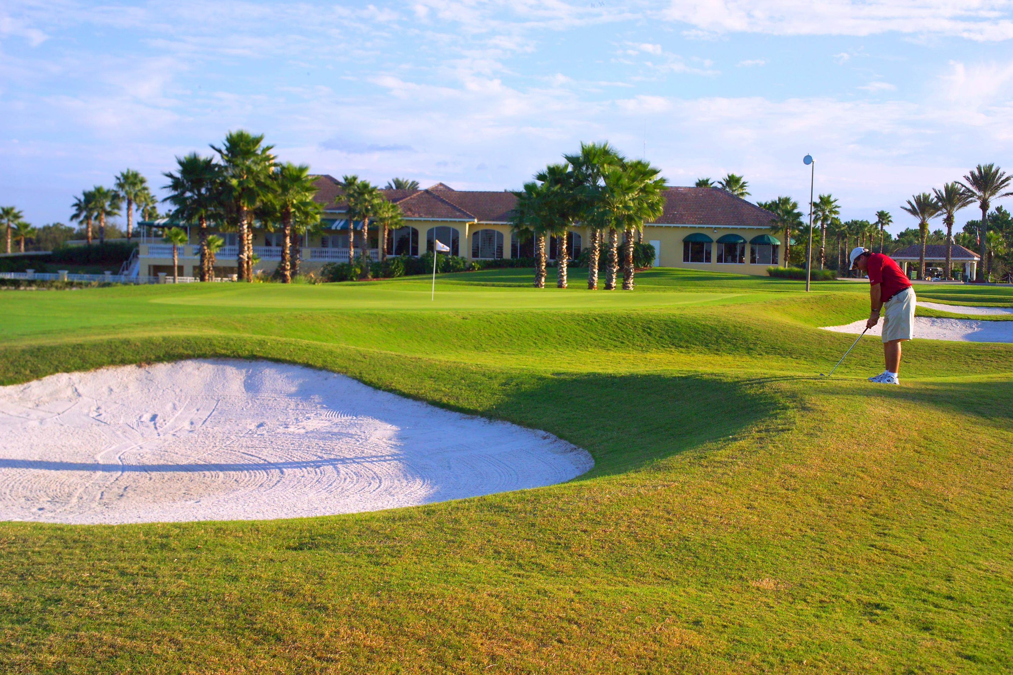 A golfer chips towards the green beside a bright white sand bunker with palm trees and the clubhouse in the background.