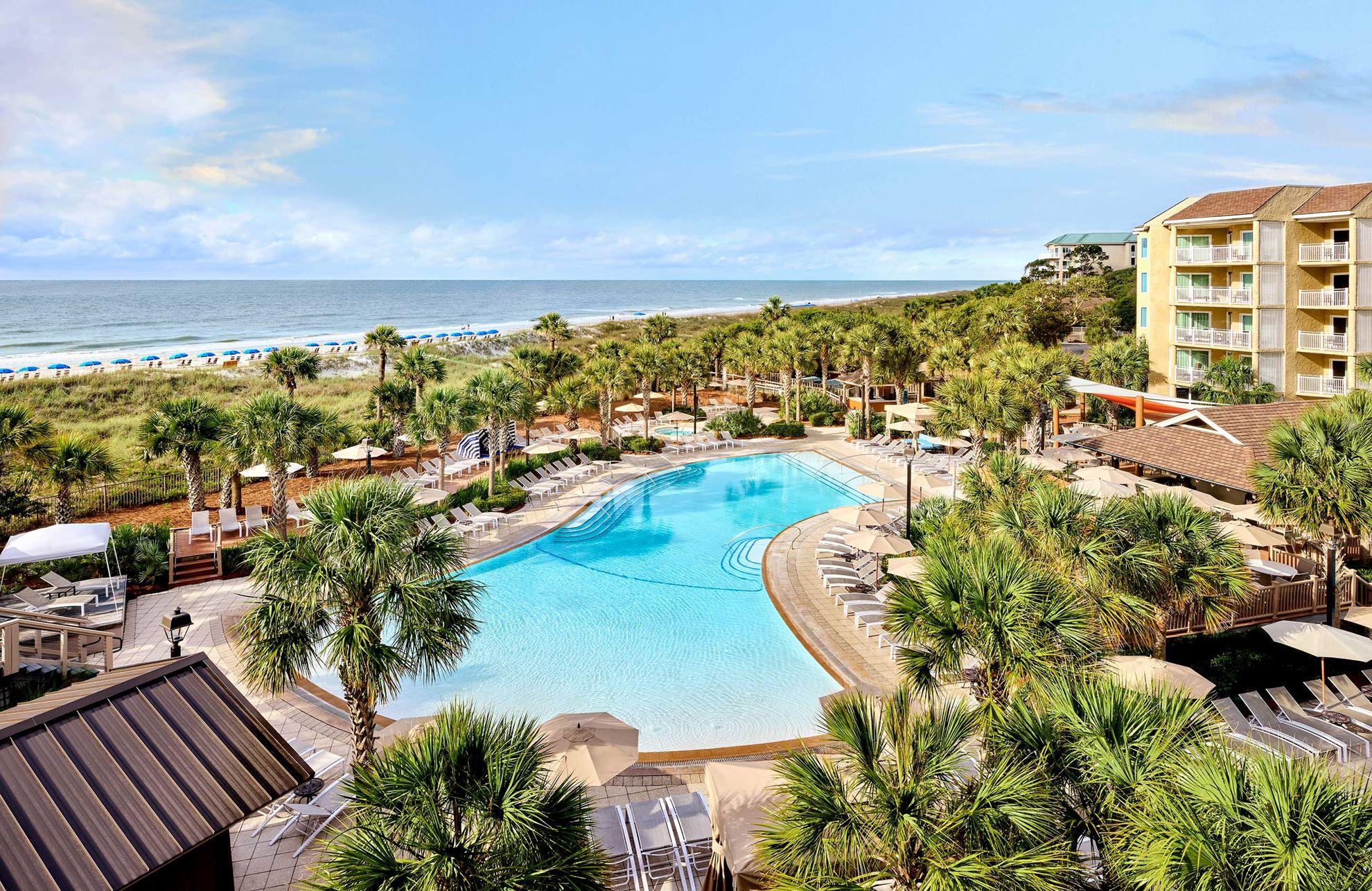 Aerial view of the swimming pool surrounded by palm trees with coastal views in the distance
