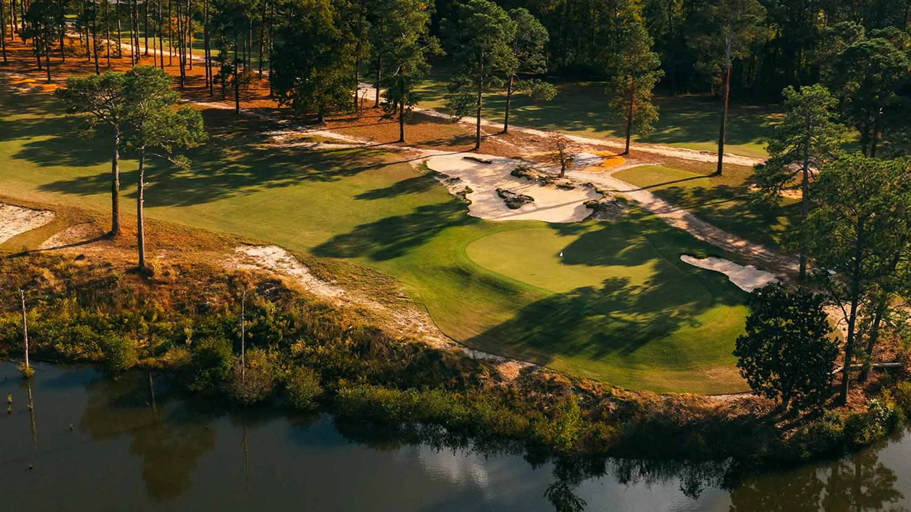 A challenging hole surrounded by sandy waste areas and towering pine trees beside a calm water hazard.