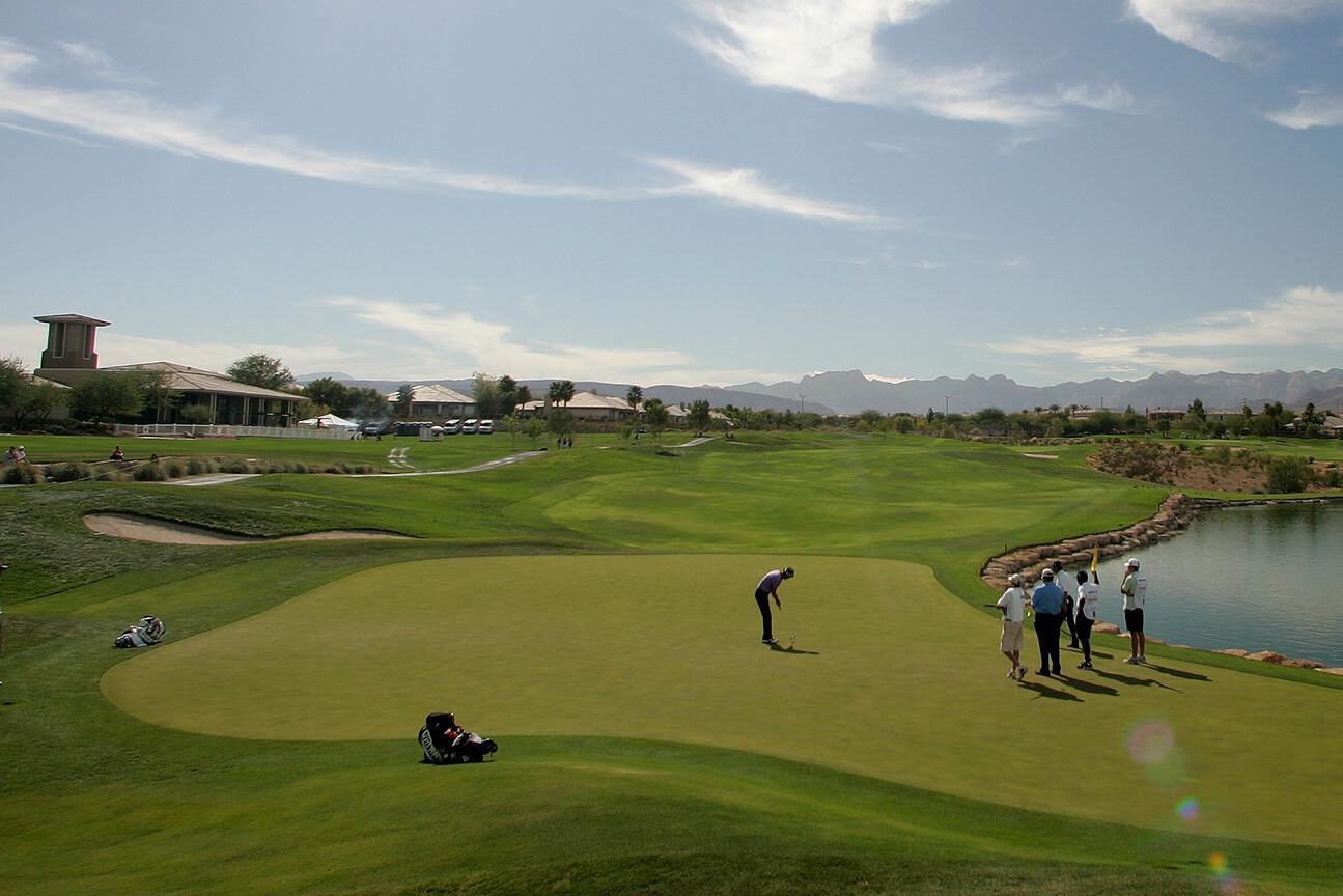 A long wide fairway leading towards golfers putting on a smooth green with mountain views in the distance