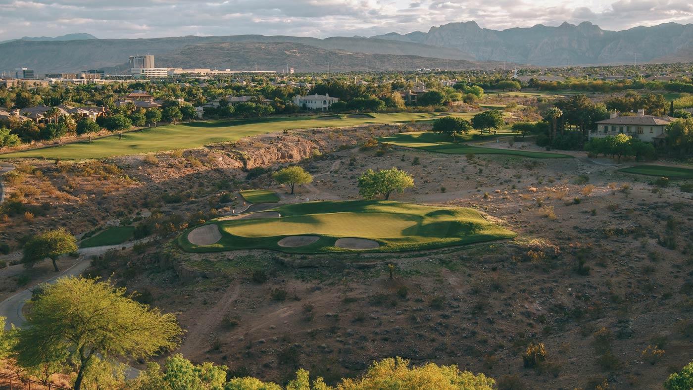 Birdseye view of an island like green littered with sand bunkers being surrounded by a rocky rough with mountains in the distance