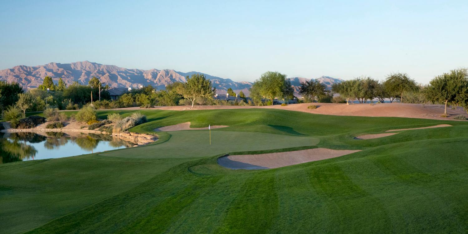 A smooth green littered with sand bunkers between rolling dunes and a water hazard reflecting the sky