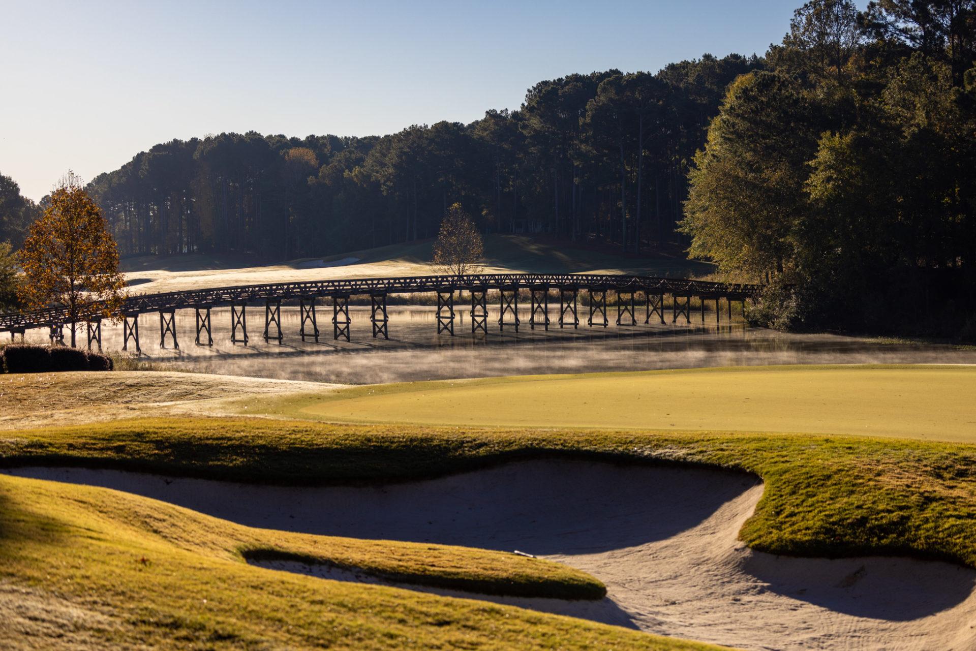 Sand bunker placed next to a smooth green with a bridge in the back to navigate the water hazard