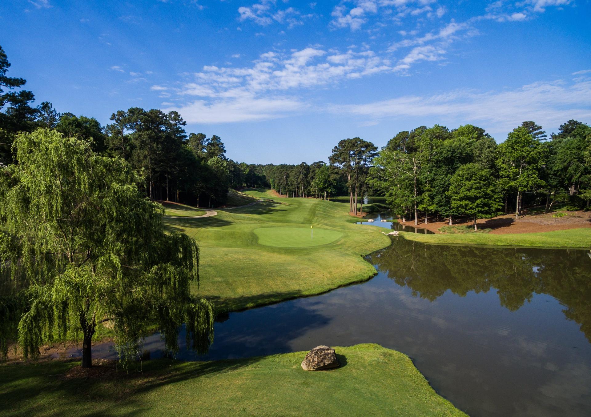 A smooth green surrounded by tall trees under cloudy blue sky's