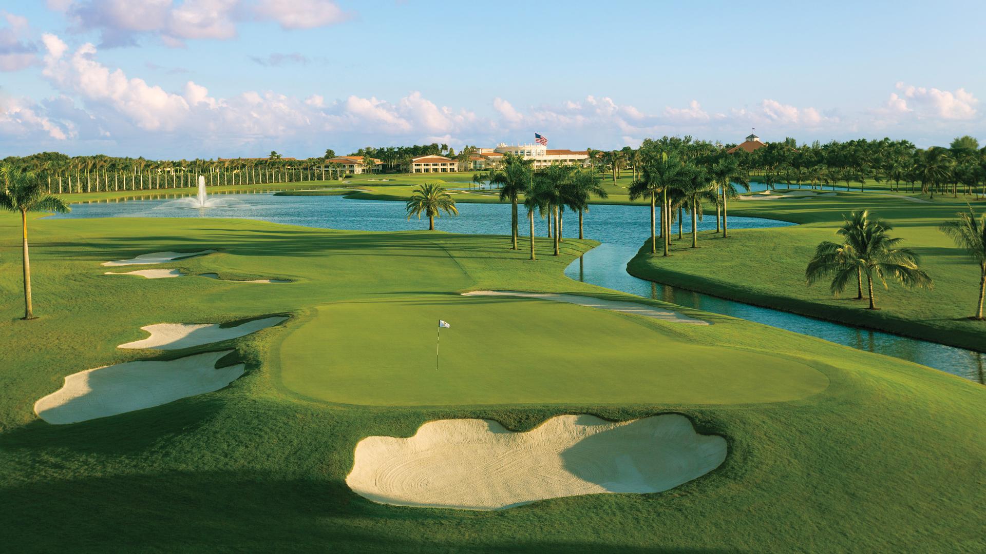 Elevated manicured green surrounded by sand bunkers and palm trees with the clubhouse in the distance