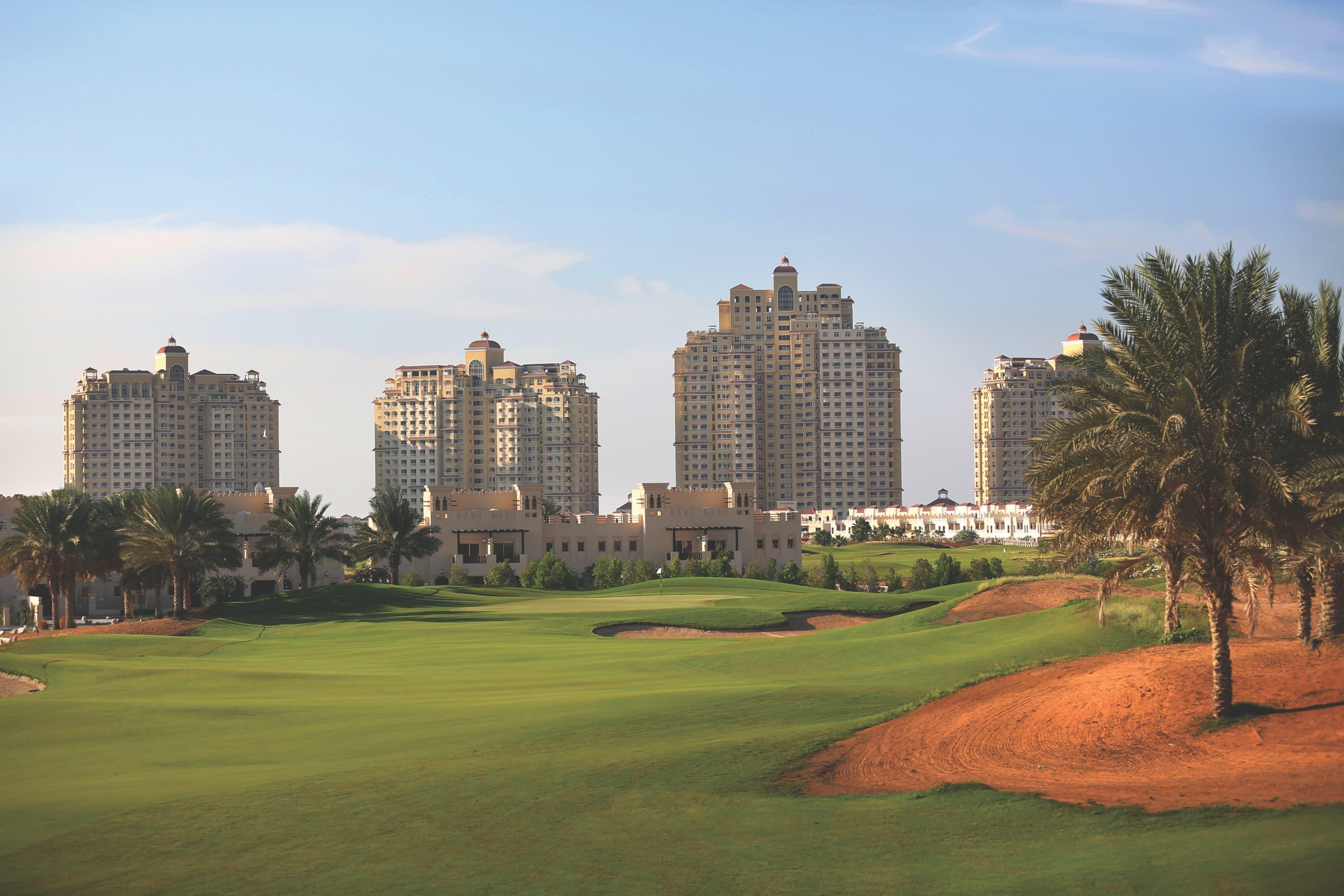 A smooth green surrounded by sand bunkers and palm trees with city buildings towering over