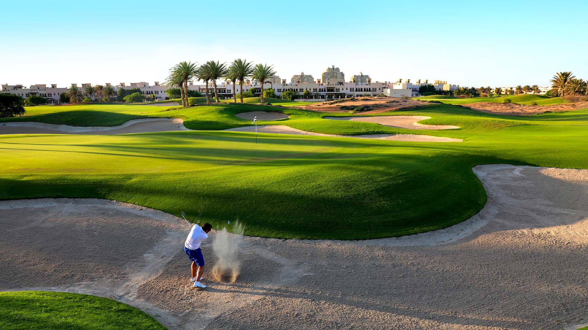 A golfer hitting out of a sand bunker towards a smooth green under blue skies