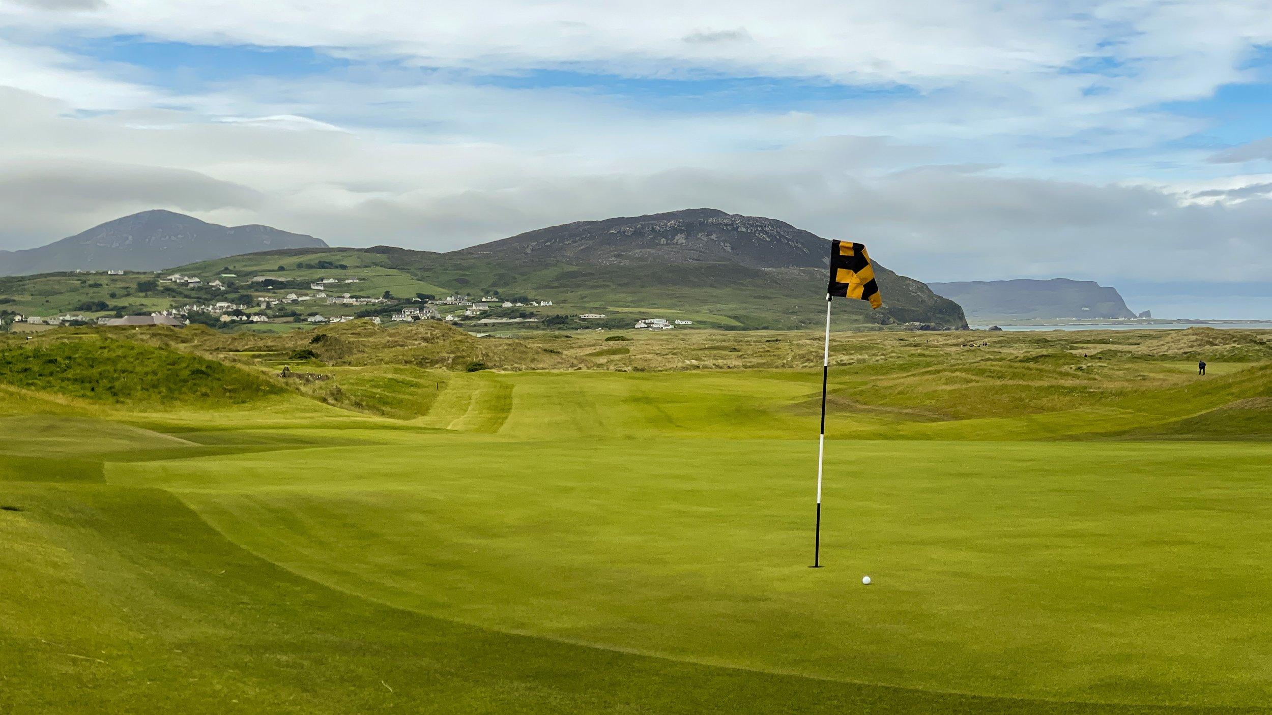Close view of a hole at the course with black and yellow flagstick and mountainous background
