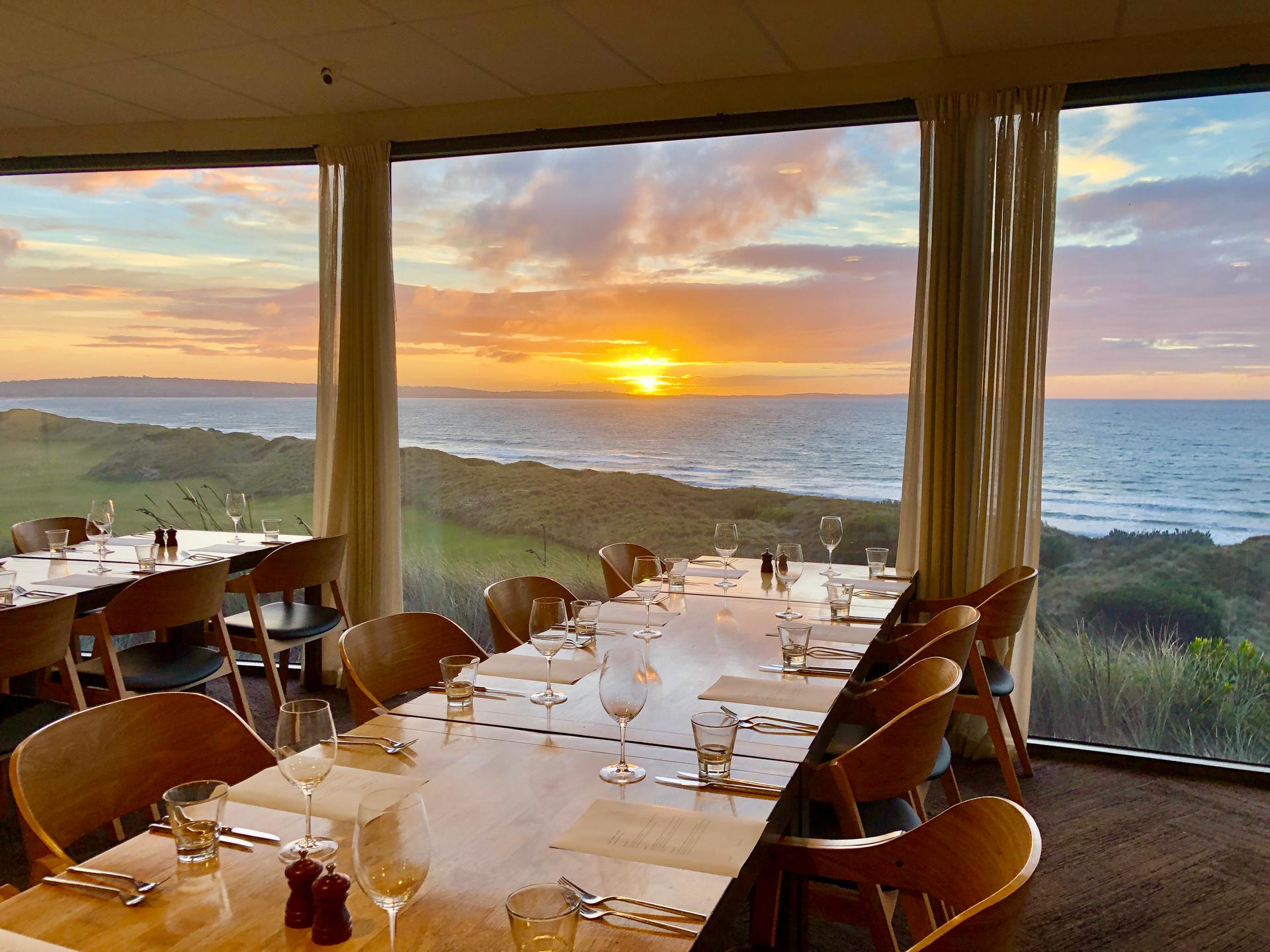 An indoor dining room with panoramic window views of the sun setting over the sea