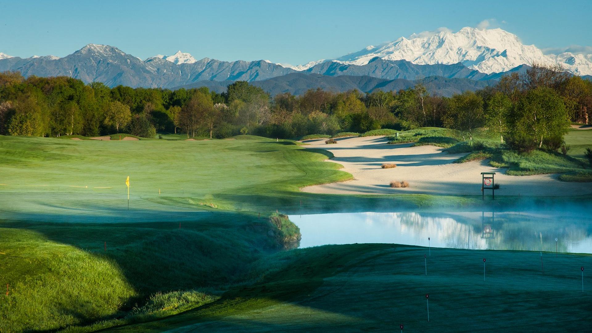 Mist rises over the course near the lake with mountains in the distance.