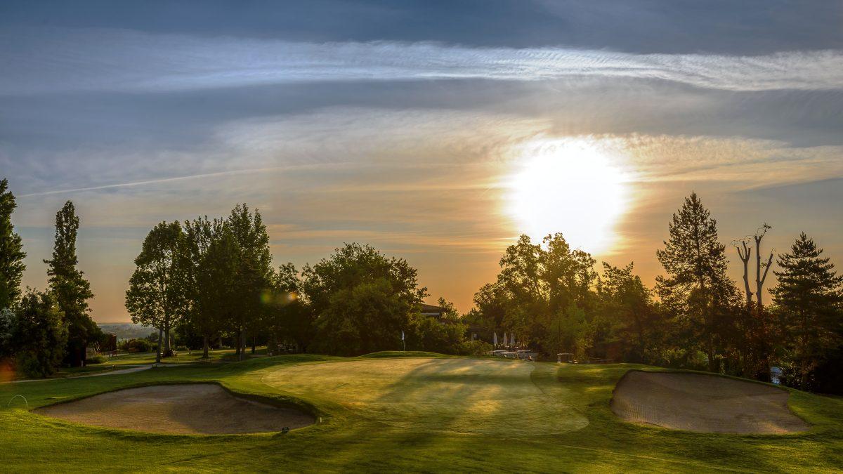 A golf course green glowing in the sunset with trees casting long shadows.