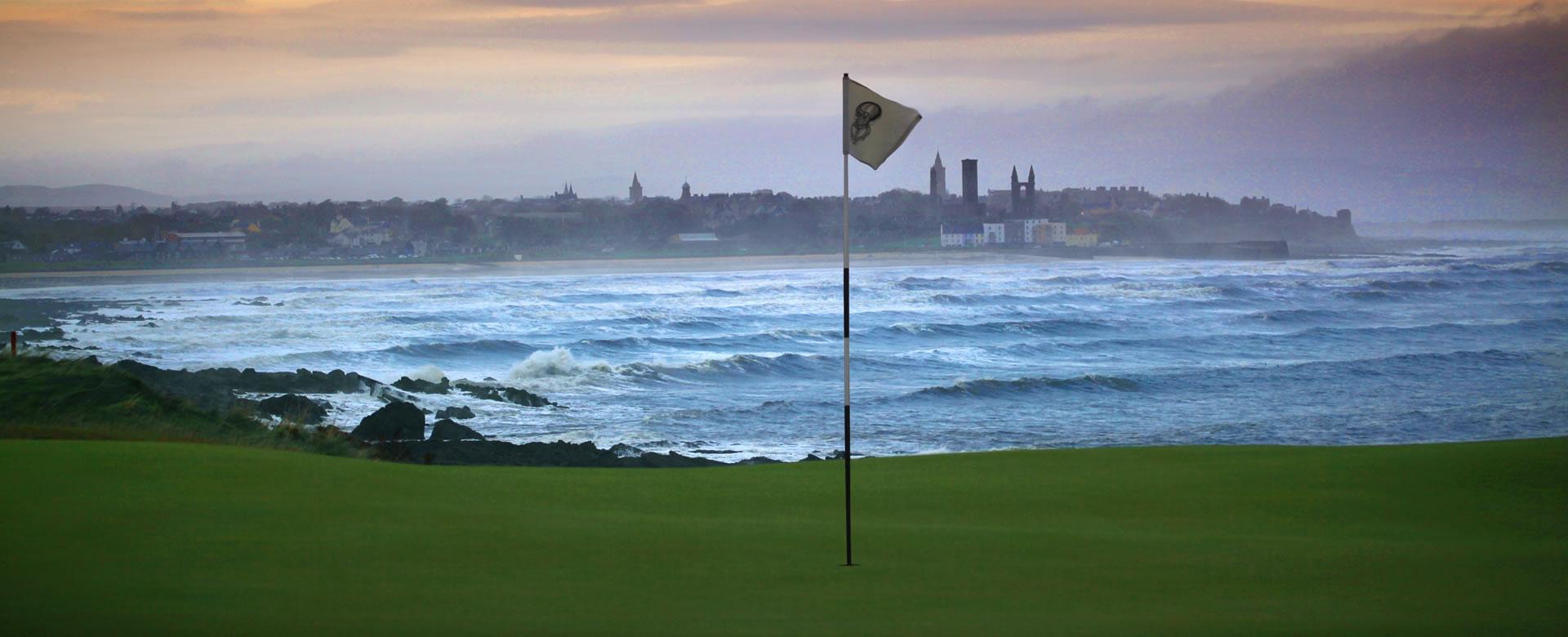 A golf flag stands tall on a cliffside green with waves crashing below and St Andrews town in the misty distance.