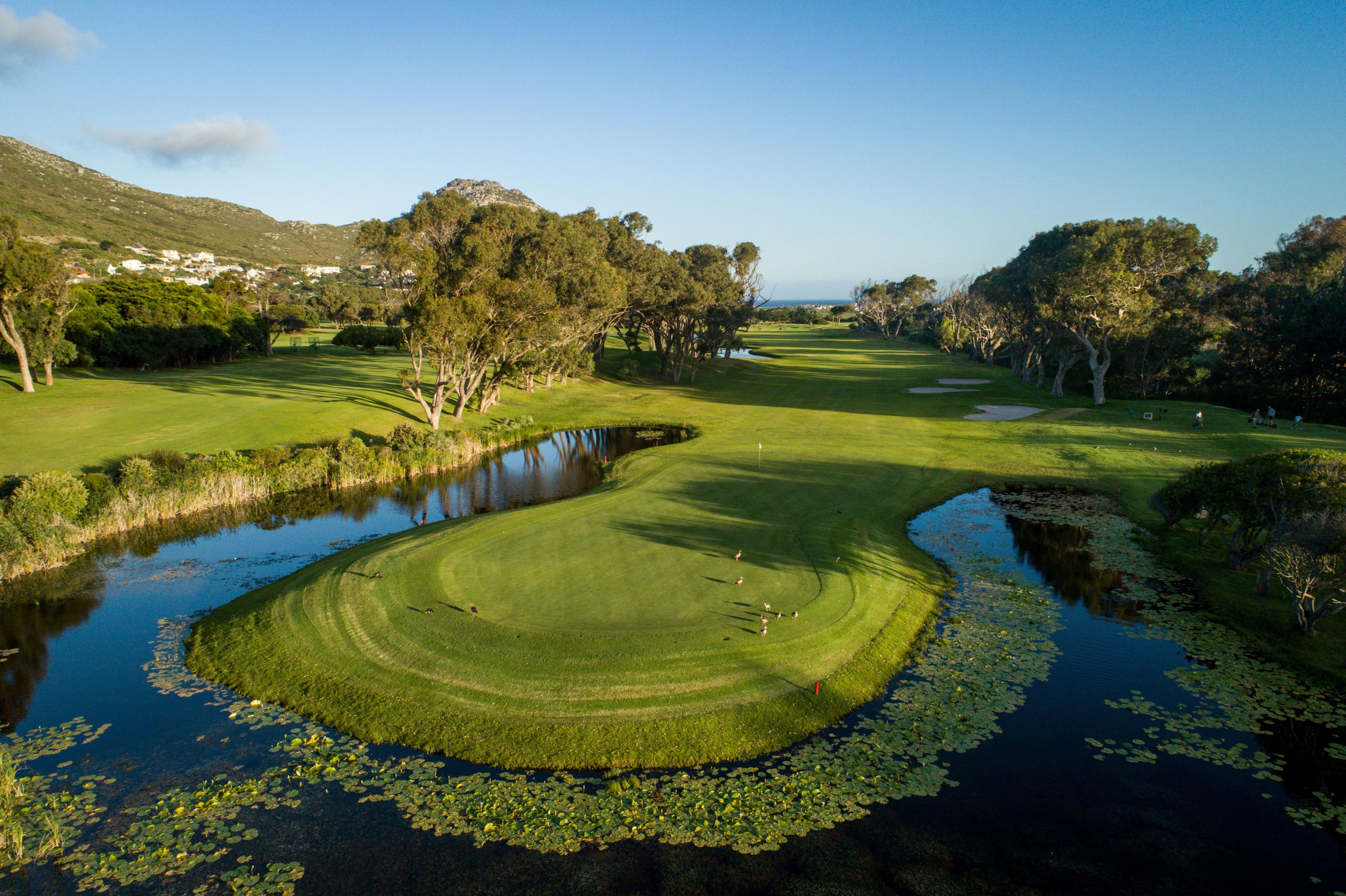 A lush green fairway surrounded by water and trees, with a peaceful mountain backdrop.