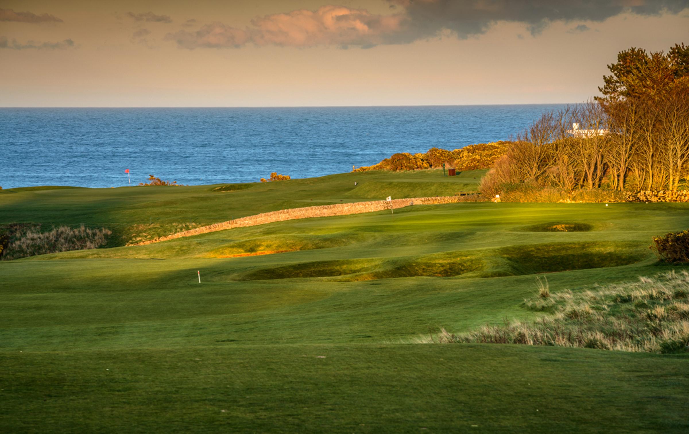 Rolling green fairways framed by stone walls and sea views at sunset.