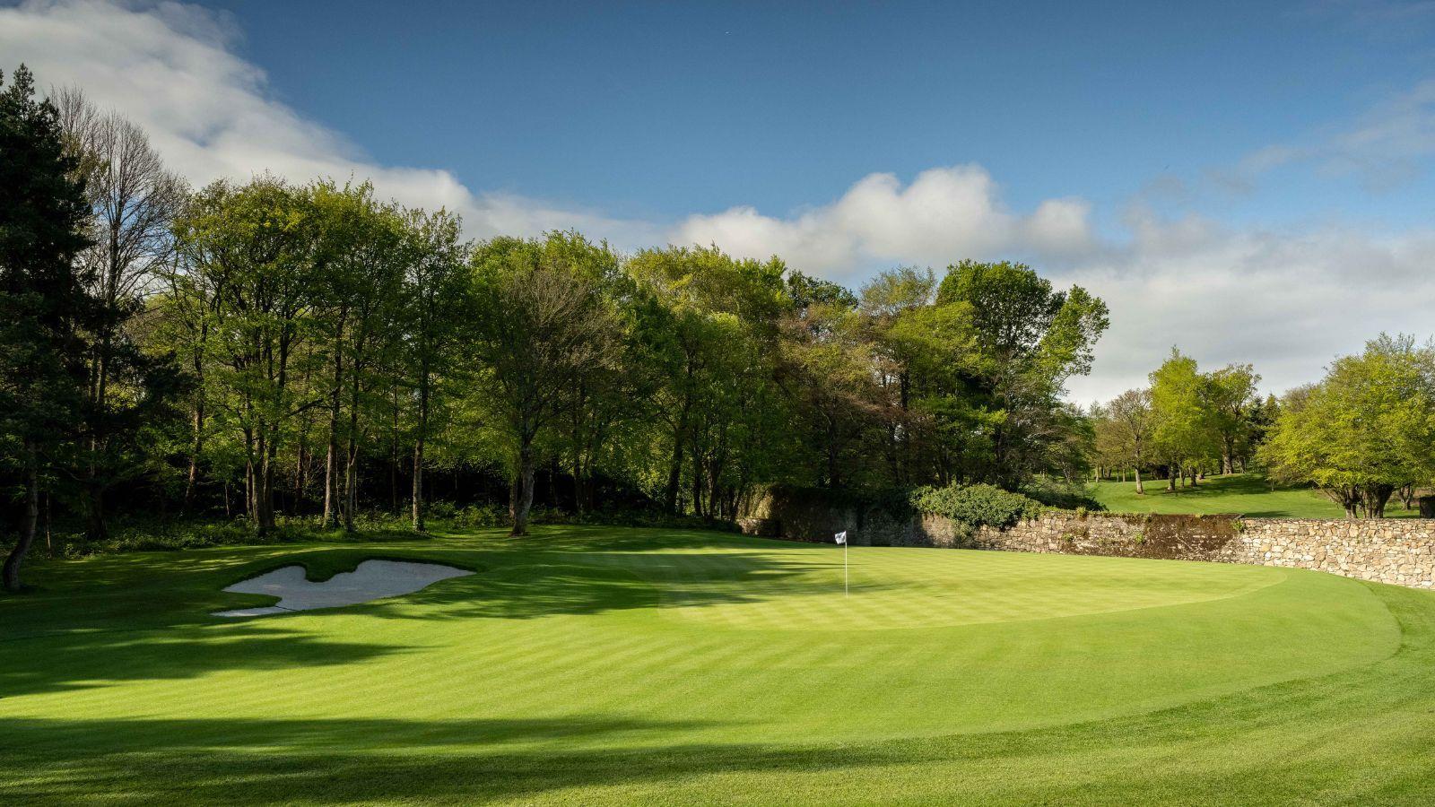 A smooth green lies next to a stone wall, with trees casting long shadows.