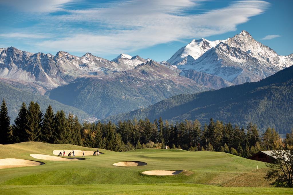 A smooth green surrounded by sand bunkers with ice top mountains in the distance