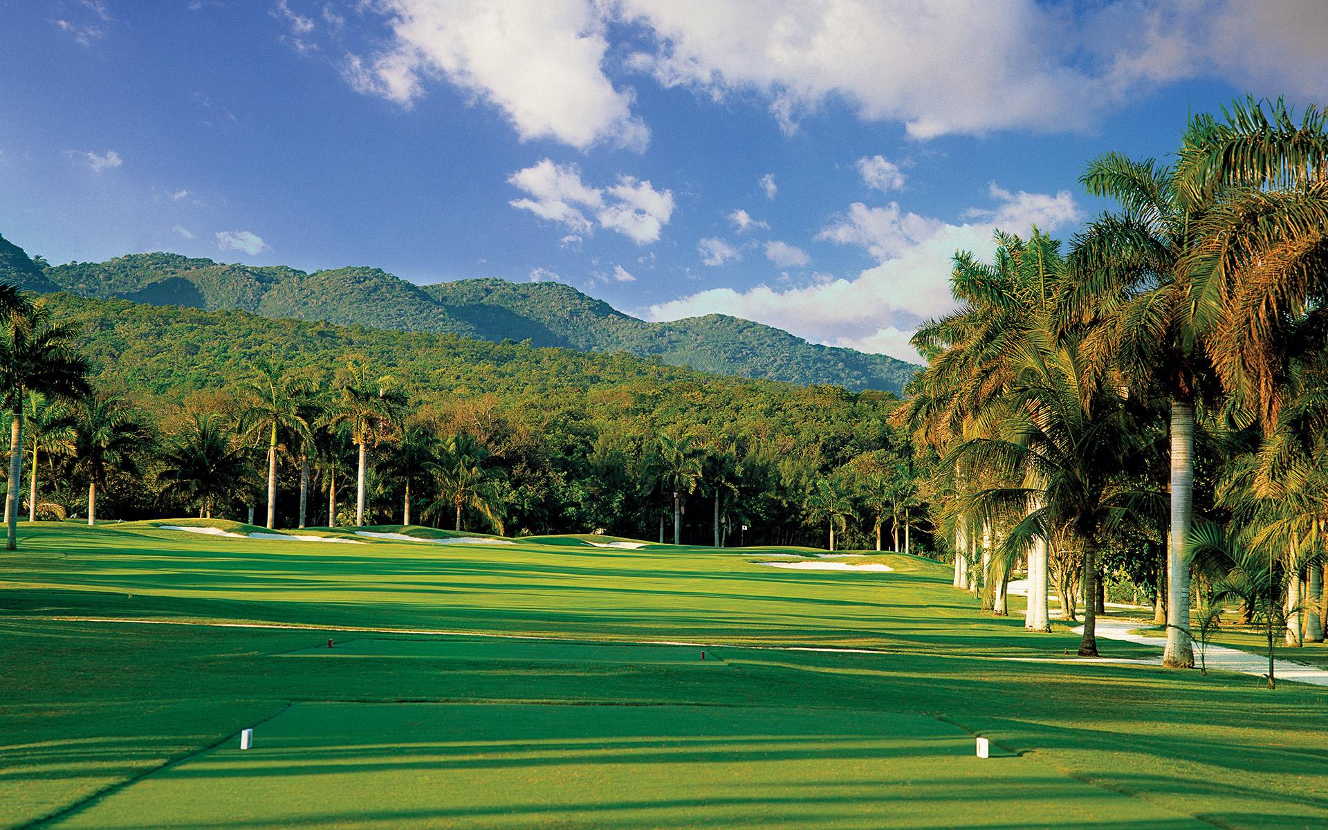 A lush tropical fairway set against a backdrop of rolling green mountains.