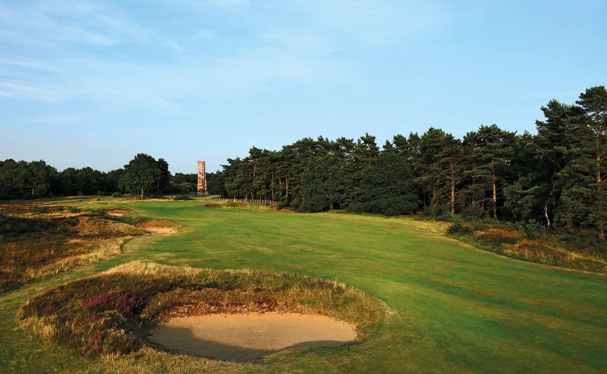 A sandy bunker sits beside a green fairway with a tower visible in the distance.