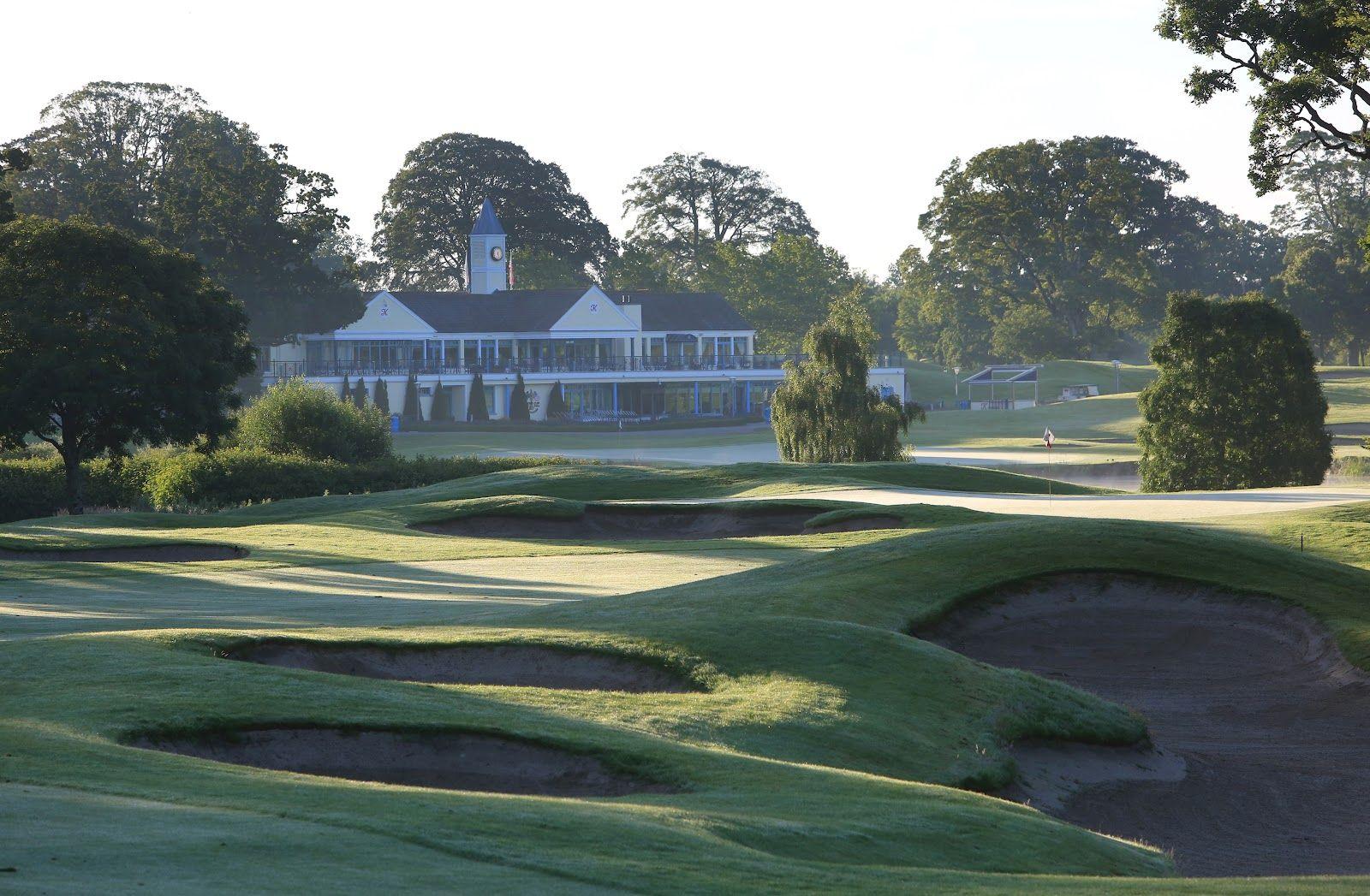 A misty morning view of a clubhouse and course with deep, undulating bunkers in the foreground.