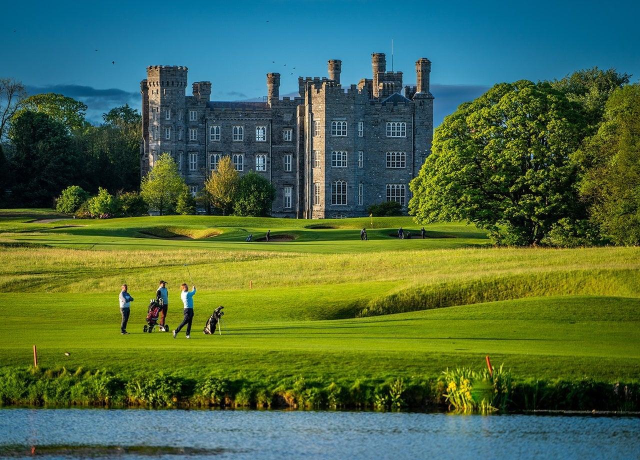 Golfers play near a castle with a pond in front.
