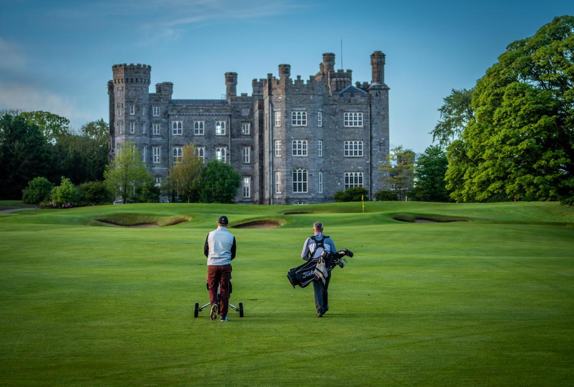 Two golfers walk toward a grand castle on lush greens.