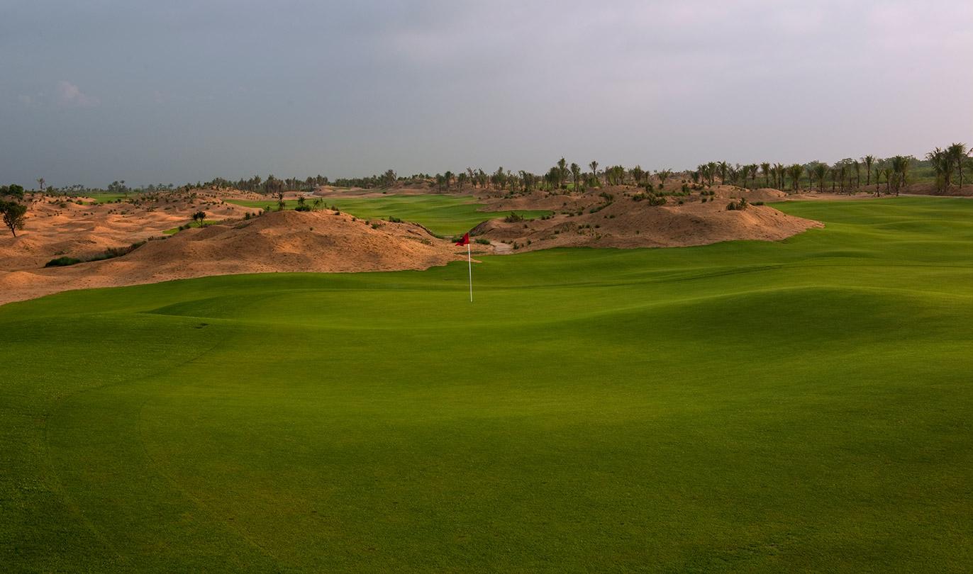 A lush green golf course surrounded by sandy desert dunes under a cloudy sky.